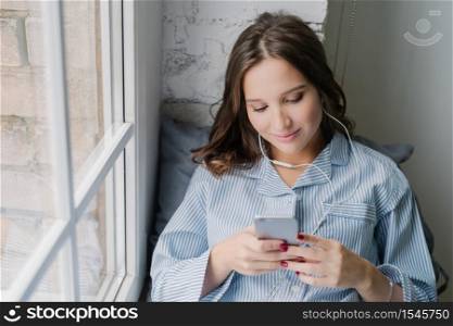 Charming woman enjoys romantic playlist, perfect sound in earphones, holds modern smart phone, dials number of boyfriend, sits on window sill, connected to wireless internet. Rest and home concept