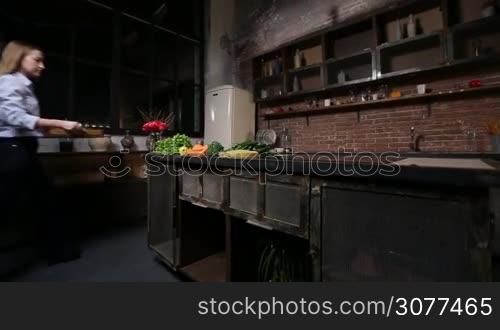 Charming woman preparing vegan smoothie with bio fruits and vegetables. Young lady bringing wooden tray with ingredients to the grunge rustic styled kitchen and laying them out on the table to prepare healthy vegetarian smoothie at home.