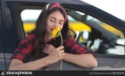 Charming young brunette woman with long brown hair smelling sunflower blossom while leaning out of car window during summer roadtrip vacation. Cheerful female enjoying leisure while travelling countryside by car.