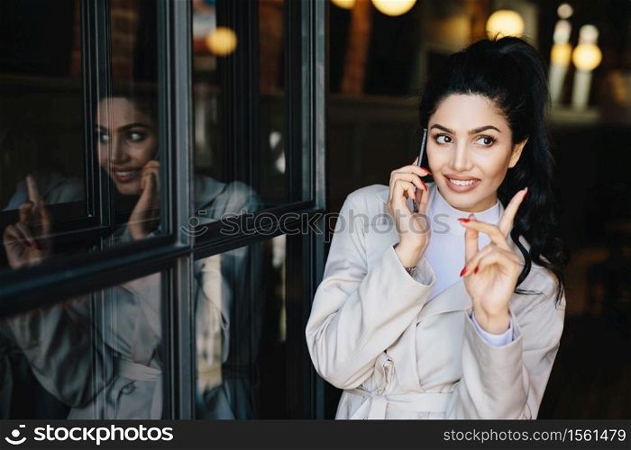Charming young brunette woman with pleasant smile having gentle hands and beautiful manicure communicating over mobile phone with her friend raising her finger while getting good idea for party