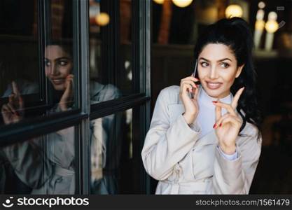 Charming young brunette woman with pleasant smile having gentle hands and beautiful manicure communicating over mobile phone with her friend raising her finger while getting good idea for party