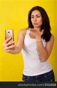 Charming young dark-haired girl in fashionable casual clothes makes a selfie on the phone and grimaces on a bright yellow background