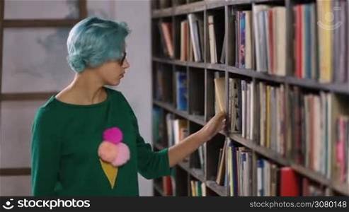 Charming young hipster woman with blue hair searching for a book in bookstore. Young cheerful woman buying books in hard cover in bookstore. Stunning female student in stylish clothes and eyeglasses selecting a book from a bookshelf.