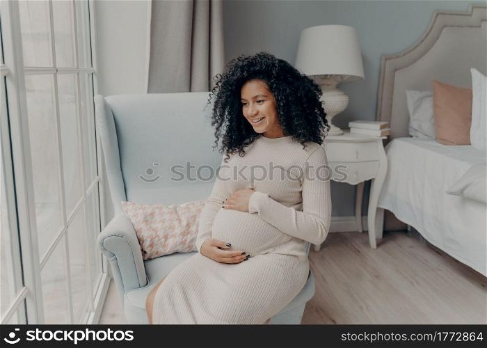 Charming young mixed race pregnant woman, future mother looking out of big window and dreaming about maternity while sitting sideways on armchair in bedroom, holding belly with both hands and smiling. Charming afro american pregnant woman looking in window and thinking about future baby