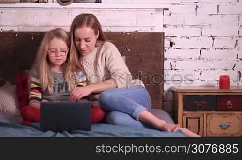 Charming young mother and lovely daughter sitting comfortably on the bed in the bedroom and eating popcorn while watching a movie on laptop together at home. Family time with movie in the evening.