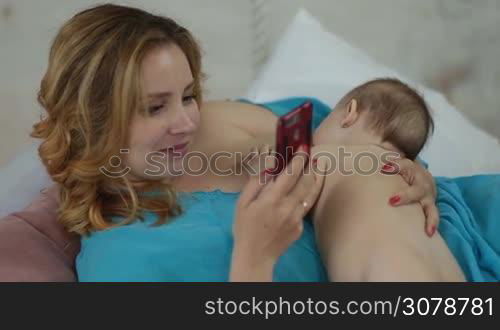 Charming young mother holding her naked baby girl and surfing the net with smartphone while breastfeeding her infant child. Portrait of attractive woman using mobile phone while feeding newborn baby at home.