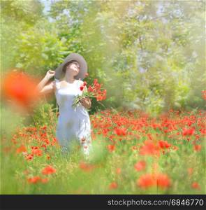 Charming young woman collects red flowers in a meadow.She is dressed in a white dress and a white hat. Small fog and sun rays