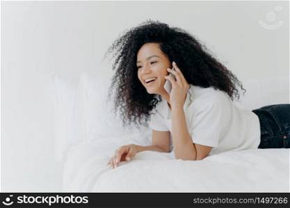 Charming young woman with crisp hair, speaks with boyfriend via smartphone, has rest in morning at bed, laughs at funny joke, dressed casually, being at home against white wall. Communication concept