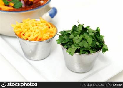 Chddar Cheese And Cilantro. Chili and beans in a ceramic bowl with onion, cilantro and cheddar cheese sprinkled on with with sides of each in stainless steel condiment cups.