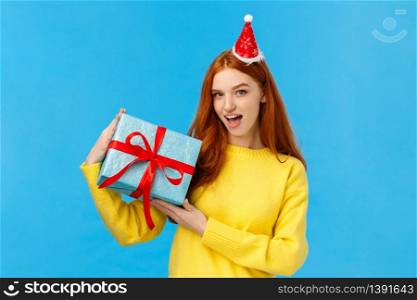 Check-out my gift. Sassy and daring good-looking redhead woman show-off, bragging with wrapped christmas present, looking camera happy and cheeky, wearing cute new year hat, blue background.. Check-out my gift. Sassy and daring good-looking redhead woman show-off, bragging with wrapped christmas present, looking camera happy and cheeky, wearing cute new year hat, blue background