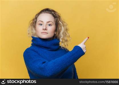 Check this out. Serious blonde young woman with curly blonde hair dressed in bright blue sweater, indicates at blank copy space for your promotional text or advertizing content. Blank yellow wall