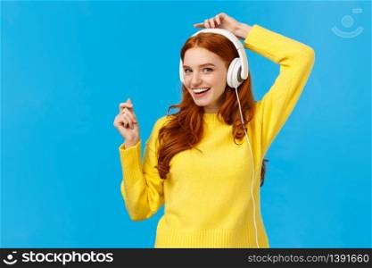 Cheeky and joyful, happy smiling redhead ecstatic woman having fun, listen music and lifting hands up as dancing, enjoying awesome sound quality, standing blue background delighted.. Cheeky and joyful, happy smiling redhead ecstatic woman having fun, listen music and lifting hands up as dancing, enjoying awesome sound quality, standing blue background delighted