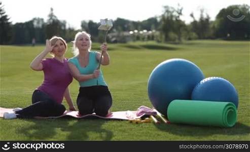Cheerful active fit adult females taking selfie with smartphone and monopod and smiling while sitting on exercise mat surrounded by sports equipment in park. Beautiful fitness senior women making selfie with mobile phone on park lawn.