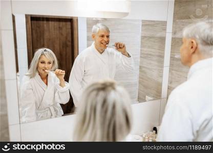 Cheerful adult couple brush teeth at the mirror in the bathroom. Grey-haired mature man and woman in bathrobes at home in the morning