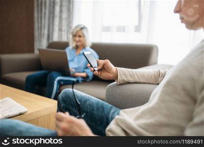 Cheerful adult love couple resting at home. Mature husband with mobile phone and wife at the laptop, happy family relax