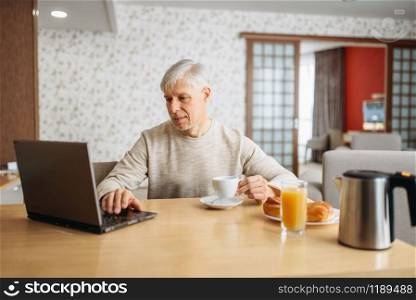 Cheerful adult man has breakfast at the laptop at home. Mature male person in jeans relax in the kitchen