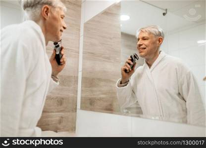 Cheerful adult man in bathrobe shaves at the mirror in the bathroom. Grey-haired mature male person with electric shaver. Cheerful adult man shaves at mirror in bathroom