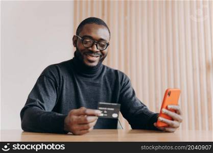 Cheerful Aframerican man entrepreneur holding credit card and using touchscreen smartphone for online shopping while making orders online sitting at home office. Ecommerce and online payment concept. Cheerful Aframerican man entrepreneur holding credit card and smartphone for online shopping