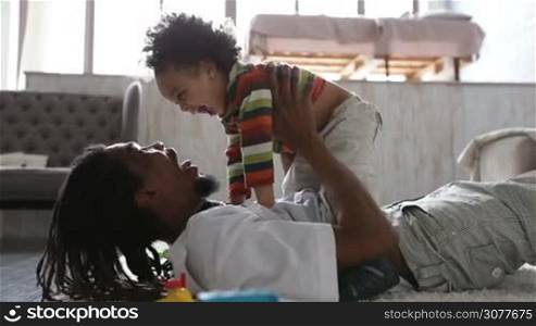 Cheerful african american father lying on the floor, holding his adorable toddler son and having fun. Smiling dad and his little child relaxing at home and fooling around while spending leisure together. Side view