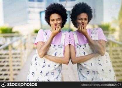 Cheerful African American female touching cheeks while making funny face and looking at camera near building with glass wall in city against blurred background. Positive black woman grimacing near building