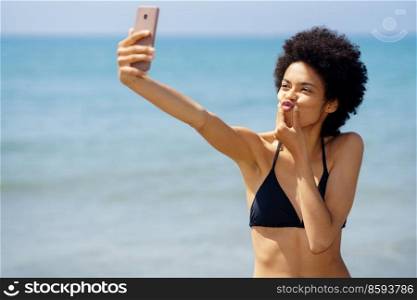 Cheerful African American female traveler in bikini pouting lips and taking self portrait on smartphone near sea in tropical resort. Funny black woman taking selfie at seaside