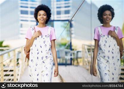 Cheerful African American woman in casual clothes walking between modern buildings smiling. Happy young female in urban background.. Cheerful African American woman in casual clothes walking between modern buildings smiling.