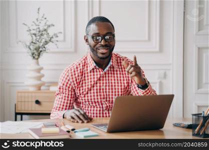 Cheerful Afro-American male English language tutor having remote classes online explains to students new topic smiling at laptop screen waving with forefinger pointing out importance of pronunciation. Cheerful Afro-American male English language tutor having remote classes online