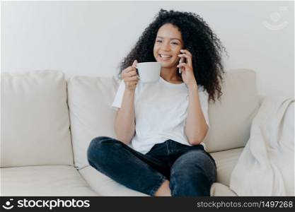 Cheerful Afro American woman has coffee break in living room, sits on couch, calls friend via modern gadget, has happy smile, thoughtful expression, discusses something pleasant. Technology, lifestyle