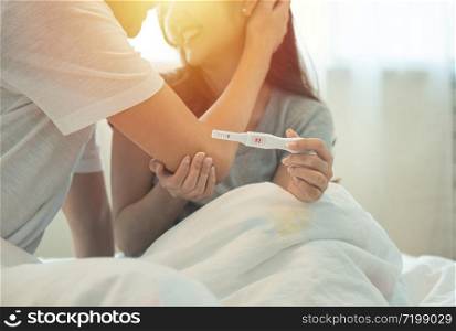 Cheerful and happy young asian man and woman with positive pregnancy test holding in hand while sitting on bed in bedroom at home.