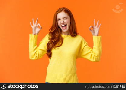 Cheerful and positive, enthusiastic redhead female in yellow sweater winking in approval, smiling joyful, give permission, positive reply, rate good movie, nod agreement, recommend, orange background.. Cheerful and positive, enthusiastic redhead female in yellow sweater winking in approval, smiling joyful, give permission, positive reply, rate good movie, nod agreement, recommend, orange background