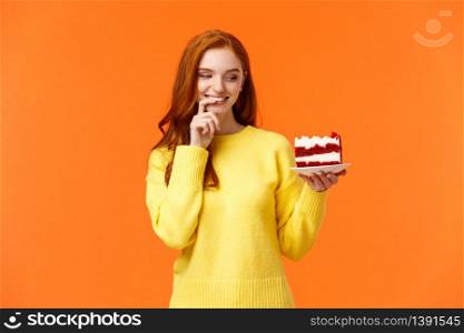 Cheerful and satisfied, happy redhead woman have cheat-day eating delicious food, holding tasty piece cake, biting lip and smiling, cant resist temptation, desire take bite, orange background.. Cheerful and satisfied, happy redhead woman have cheat-day eating delicious food, holding tasty piece cake, biting lip and smiling, cant resist temptation, desire take bite, orange background