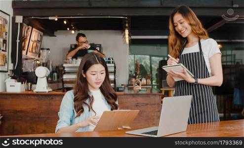 Cheerful Asian lady waitress with notebook taking receive order menu from young client girl at urban cafe. Young Asian freelance women working on laptop at coffee shop. Owner small business concept.