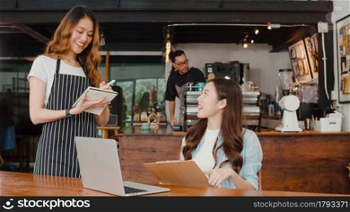 Cheerful Asian lady waitress with notebook taking receive order menu from young client girl at urban cafe. Young Asian freelance women working on laptop at coffee shop. Owner small business concept.