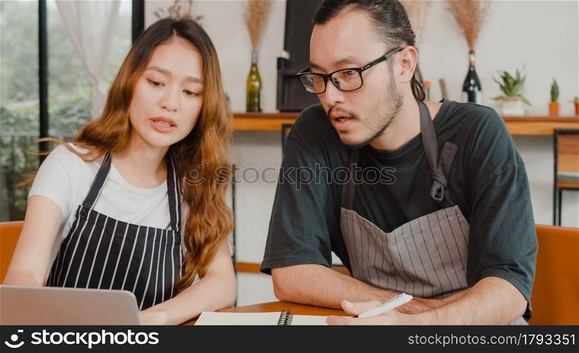 Cheerful Asian small business owners couple using laptop discussing project and finance with documents of shop for the month at cafe. Woman and man doing paperwork together, paying taxes online.