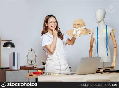 Cheerful Asian young female designer wearing eyeglasses doing fashion design on beautiful sombrero in studio, full of tailoring tools  with mannequin and equipment on the desk