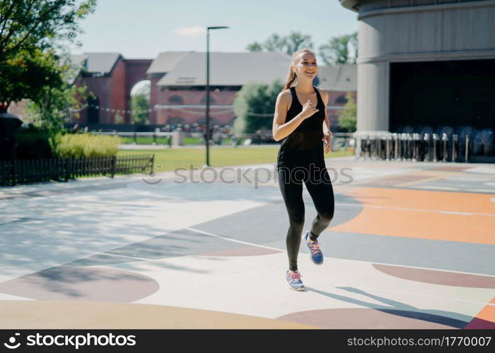 Cheerful athletic woman runs actively on stadium dressed in black sportclothes enjoys physical activities outdoors during summer day being full of energy. People sport and motivation concept