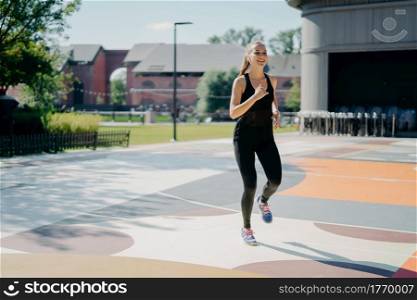 Cheerful athletic woman runs actively on stadium dressed in black sportclothes enjoys physical activities outdoors during summer day being full of energy. People sport and motivation concept