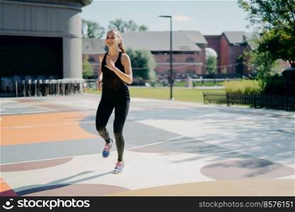 Cheerful athletic woman runs actively on stadium dressed in black sportclothes enjoys physical activities outdoors during summer day being full of energy. People sport and motivation concept
