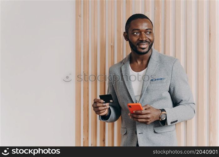 Cheerful attractive Aframerican man entrepreneur in stylish blazer holding credit card and smartphone in hands, making payment online or shopping via mobile Internet. Technology and e-commerce concept. Cheerful Aframerican man entrepreneur in stylish blazer holding credit card and smartphone in hands