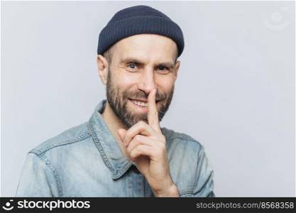 Cheerful attractive male with blue eyes shows silence sign, has satisfied expression, wears fashionable hat and denim jacket, isolated over grey background. Handsome man shows hush sign. Be quiet 
