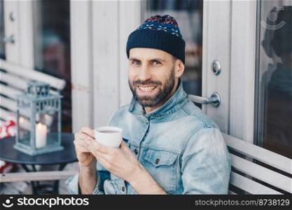 Cheerful bearded male in denim jacket, has positive happy expression, spends free time at terrace, drinks aromatic tea, feels relaxed and delighted. People, positive emotions and recreation time