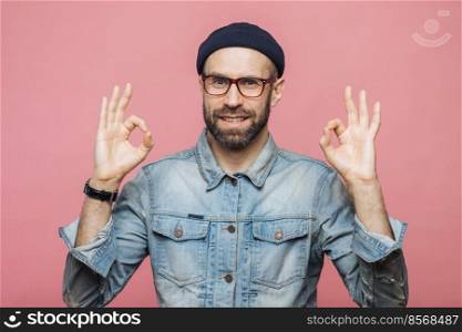 Cheerful bearded male in fashionable clothing, makes ok sign, being satisfied with something, isolated over pink studio background. Unshaven middle aged man gestures indoor, shows his approval