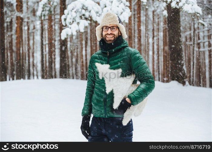 Cheerful bearded male wears warm anorak and hat, walks in winter forest, holds artificial fir tree, has festive mood, going to celebrate New Year. Outdoor shot of male spends free time on cold weather