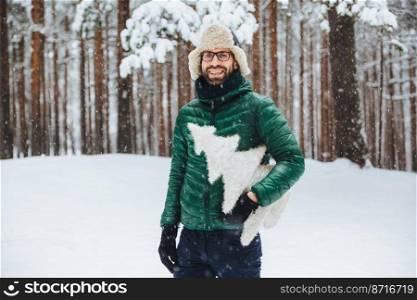 Cheerful bearded male wears warm anorak and hat, walks in winter forest, holds artificial fir tree, has festive mood, going to celebrate New Year. Outdoor shot of male spends free time on cold weather