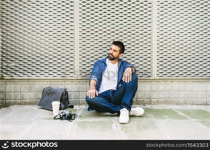 Cheerful bearded man in casual attire sitting on street ground while relaxing on a coffee break