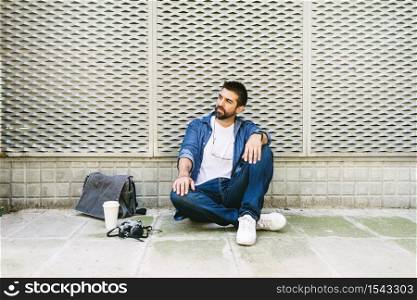 Cheerful bearded man in casual attire sitting on street ground while relaxing on a coffee break