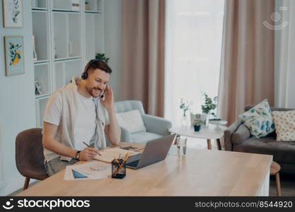 Cheerful bearded young man wearing headphones having online meeting making video call on laptop while working at cozy workplace at home office, sitting in modern living room Remote job concept. Happy guy making notes in notebook during online lesson at home
