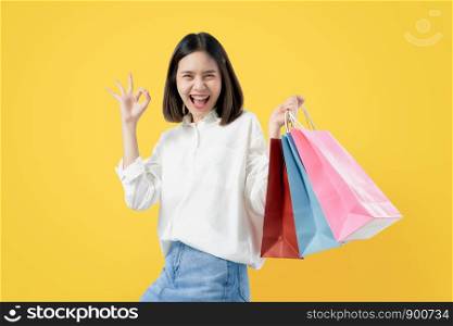 Cheerful beautiful Asian woman holding multi coloured shopping bags and shows ok sign on light yellow background.