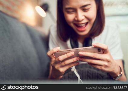 Cheerful beautiful Asian woman holding smartphone and playing games mobile with headphones on sofa in house.