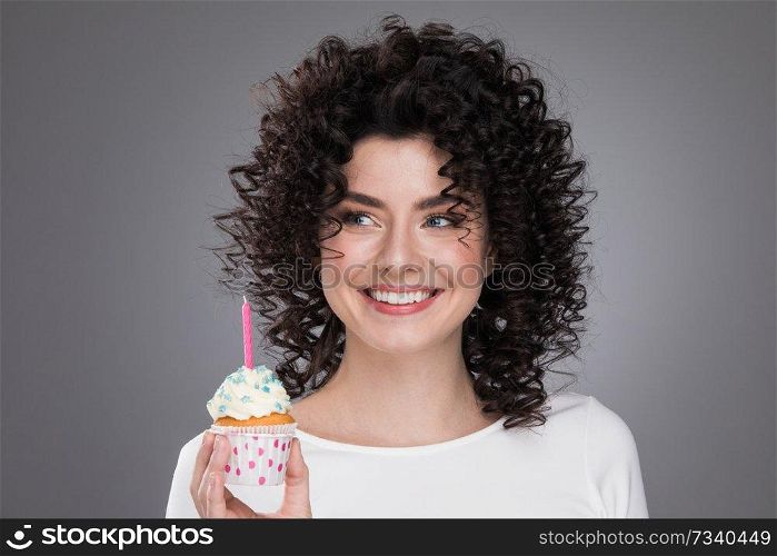 Cheerful beautiful curly young woman making a wish holding birthday cupcake with candle. Woman with birthday cupcake